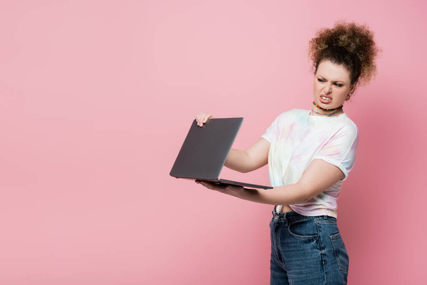 Disgusted curly woman holding laptop isolated on pink  - Foto, immagini
