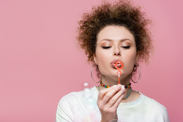 Curly woman blowing soap bubbles isolated on pink  - Fotó, kép