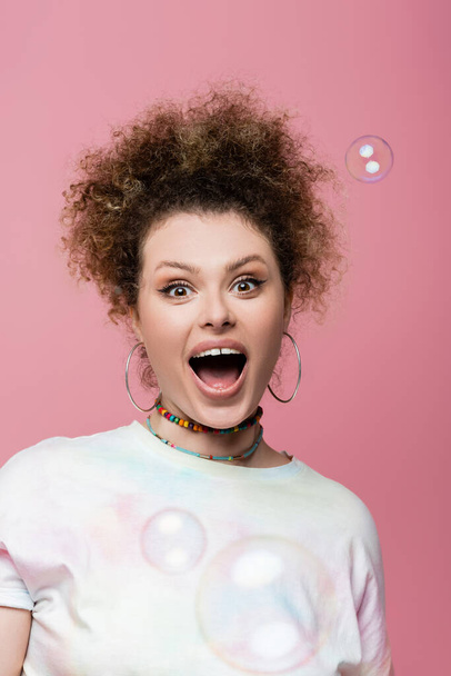 Excited curly woman looking at camera near soap bubbles on pink background - Фото, изображение