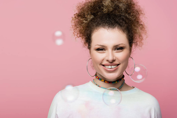 Pretty curly woman looking at camera near soap bubbles on pink background - Foto, Bild