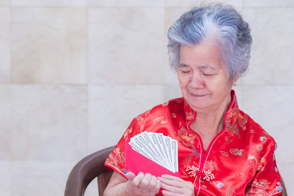 Elderly Asian woman wearing a Cheongsam red shirt, holding a red envelope packet with US dollars (or Ang Pao) for giving to children during Chinese New Year. Selective focus. Concept of festival. - Photo, Image
