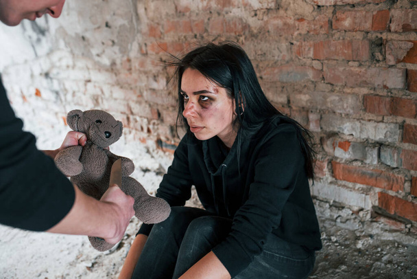 Hombre violento de pie sosteniendo oso de peluche en las manos y amenaza a la chica que se sienta en el suelo en un edificio abandonado. - Foto, Imagen
