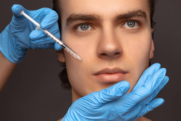 Close-up face of young man and female hands in blue gloves with syringe isolated on dark background. Cosmetic, filling surgery procedures. Anti-aging concept. - Foto, imagen