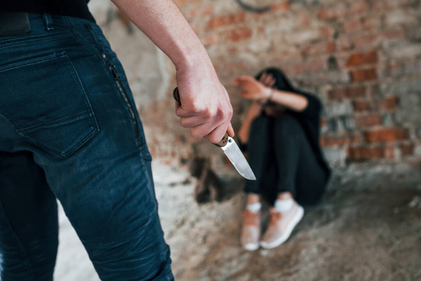 Violent man standing with knife in hand and threatens girl that sits on the floor in abandoned building. - Foto, Bild