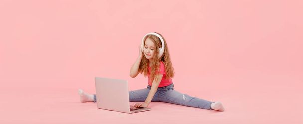 чарівна дівчинка сидить у шпагаті поруч з ноутбуком. дитина в білих навушниках розтягується і вивчає домашнє завдання або дивиться мультфільми, слухаючи музику
. - Фото, зображення