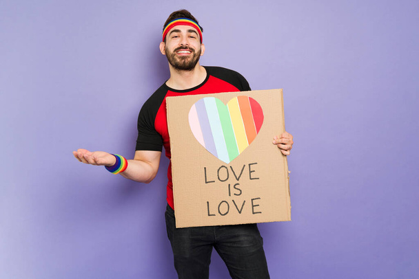 Studio shot of a cheerful gay man smiling while showing a support sign with a love is love message - Foto, immagini