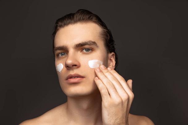Facial skin care for man. Young happy man with perfect well-kept skin using face cream isolated over grey background. Cosmetics, health care, skin care, beauty - Photo, Image