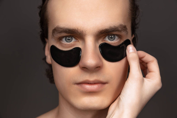 Young adorable man with well-kept skin using patches under eyes isolated over grey background. Fashion, cosmetics, health care, skin care, beauty - 写真・画像