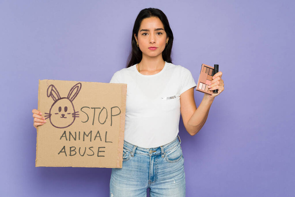Upset young woman holding a protest sign with a stop animal cruelty message in the cosmetic industry - 写真・画像