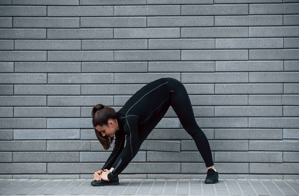 Young sportive girl in black sportswear outdoors doing stretching near gray wall. - Photo, Image