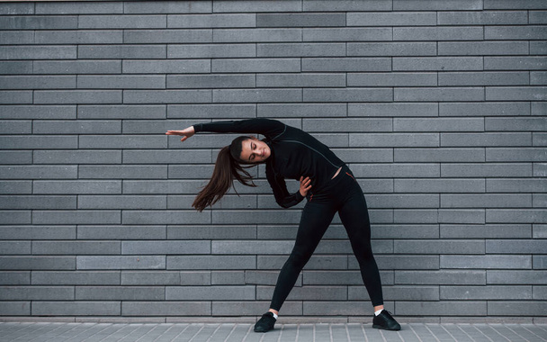 Jovem menina esportiva em sportswear preto ao ar livre fazendo alongamento perto da parede cinza. - Foto, Imagem