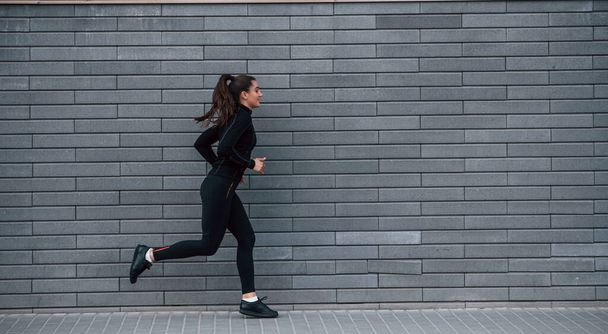 Young sportive girl in black sportswear running outdoors near gray wall. - Photo, Image