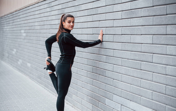 Young sportive girl in black sportswear outdoors doing stretching near gray wall. - Фото, изображение