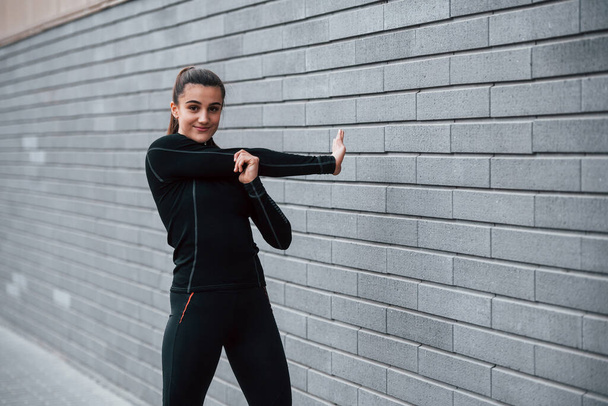 jong sportief meisje in zwart sportkleding buiten doen stretching buurt grijs muur. - Foto, afbeelding