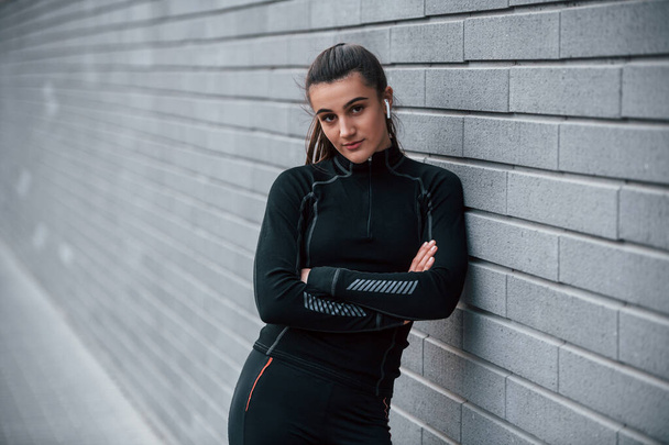 Young sportive girl in black sportswear outdoors near gray wall. - 写真・画像