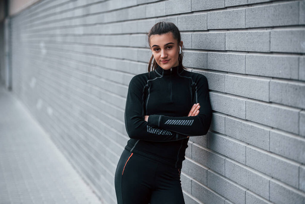 Young sportive girl in black sportswear outdoors near gray wall. - Фото, изображение