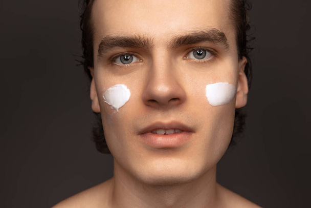Facial skin care for man. Young happy man with perfect well-kept skin using face cream isolated over grey background. Cosmetics, health care, skin care, beauty - Фото, зображення