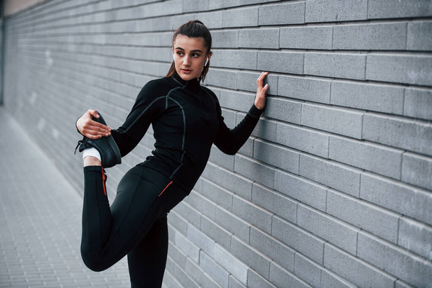 Young sportive girl in black sportswear outdoors doing stretching near gray wall. - Foto, Imagem