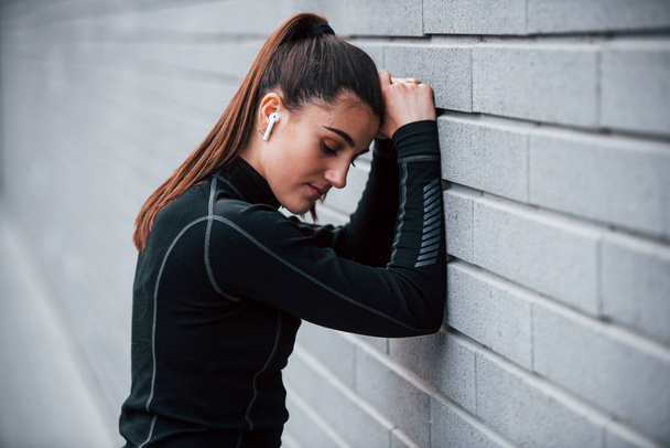 Young sportive girl in black sportswear outdoors near gray wall. - Фото, зображення