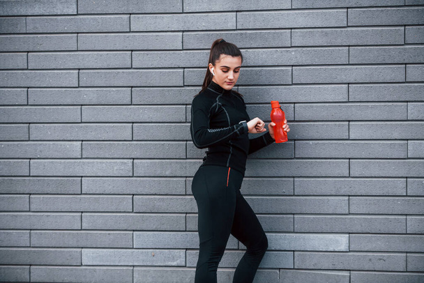 Młoda sportowa dziewczyna w czarnej odzieży sportowej stojąca na zewnątrz przy szarej ścianie z butelką wody i robiąca sobie przerwę. - Zdjęcie, obraz
