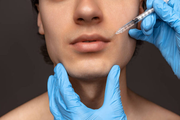 Close-up face of young man and female hands in blue gloves with syringe isolated on dark background. Cosmetic, filling surgery procedures. Anti-aging concept. - Фото, изображение