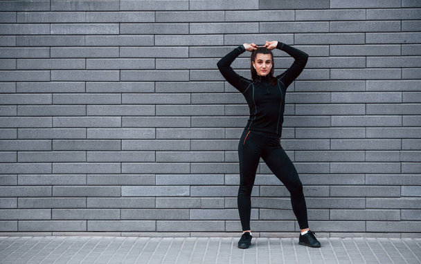 Young sportive girl in black sportswear standing outdoors near gray wall. - Foto, afbeelding