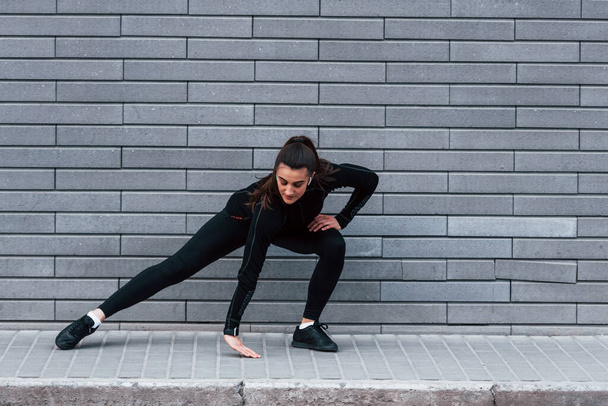 Young sportive girl in black sportswear outdoors doing stretching near gray wall. - 写真・画像