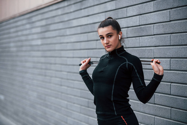 Young sportive girl in black sportswear standing with jump rope in hands outdoors near gray wall. - Foto, Imagem