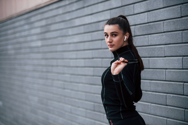 Young sportive girl in black sportswear standing with jump rope in hands outdoors near gray wall. - Foto, immagini