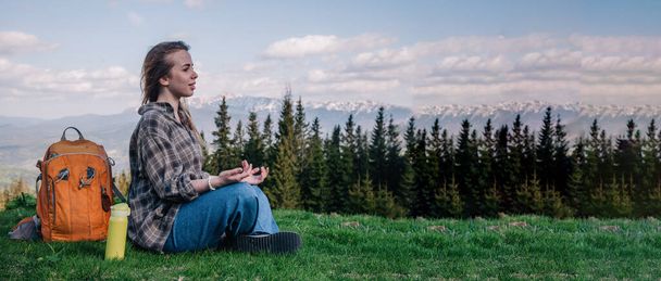 A young, slender girl in a plaid shirt and jeans with an orange backpack meditates sitting on the green grass against the backdrop of mountains. photo on horizontal banner - Zdjęcie, obraz