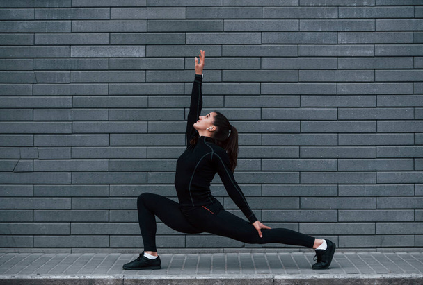 Young sportive girl in black sportswear outdoors doing yoga exercises near gray wall. - Φωτογραφία, εικόνα