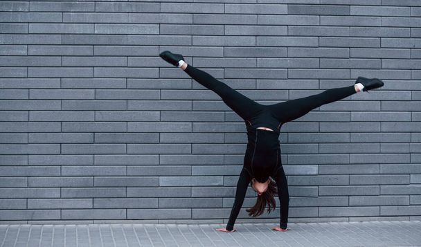 Young sportive girl in black sportswear doing hard handstand exercises outdoors near gray wall. - Фото, зображення
