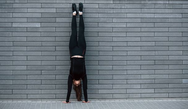 Young sportive girl in black sportswear doing hard handstand exercises outdoors near gray wall. - Φωτογραφία, εικόνα