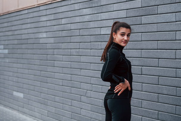 Young sportive girl in black sportswear standing outdoors near gray wall. - Photo, image
