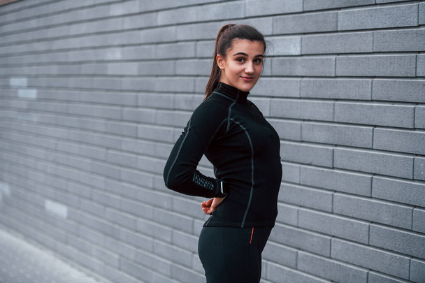Young sportive girl in black sportswear standing outdoors near gray wall. - Foto, Bild
