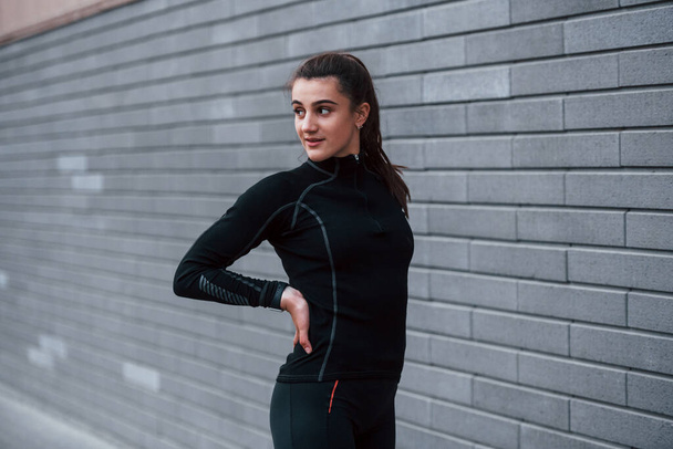 Young sportive girl in black sportswear standing outdoors near gray wall. - Foto, imagen