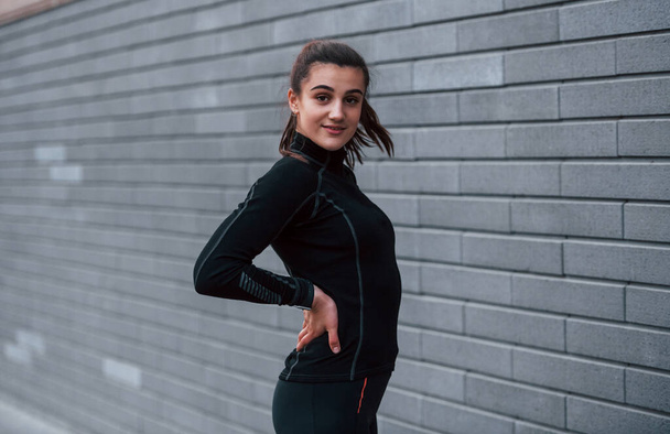 Young sportive girl in black sportswear standing outdoors near gray wall. - Foto, Bild