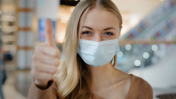 Indoors portrait caucasian woman patient girl female lady in face protective medical mask protecting health from coronavirus infection covid shows like puts thumbs up recommends wearing respirator - Foto, Imagen