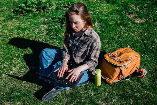 A young, slender girl with loose hair in a plaid shirt and jeans with an orange backpack works on a laptop sitting on green grass against the backdrop of mountains. view from above - Photo, image