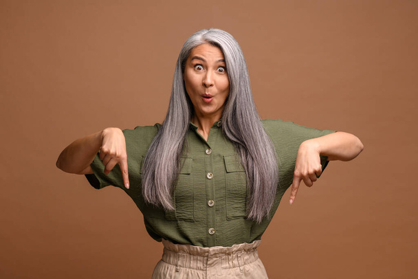 Cheerful positive woman pointing fingers down showing space for your advertisement, looking at camera with surprised face. Indoor studio shot isolated on brown - Foto, Imagen