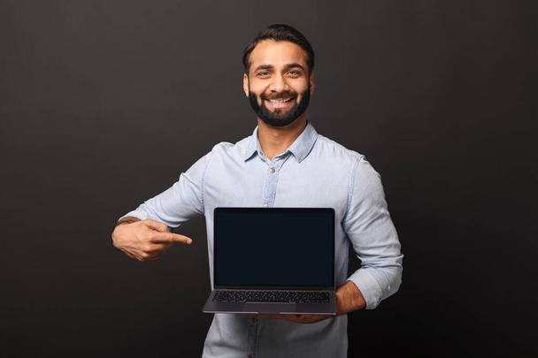 Happy Indian man holding laptop with empty screen and points finger at it - Valokuva, kuva