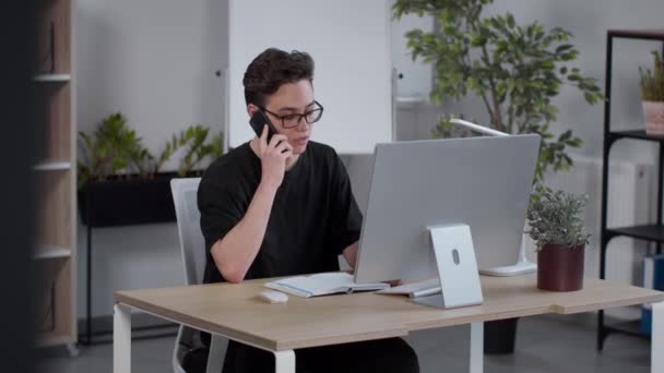 Młody utalentowany biznesmen noszący okulary konsultant klienta przez smartfona, pracujący online na komputerze w biurze - Materiał filmowy, wideo