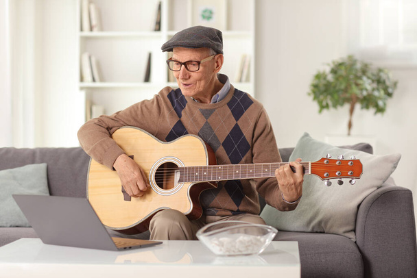 Anciano tocando una guitarra acústica y sentado frente a una computadora portátil en casa  - Foto, Imagen