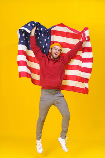 Cheerful young man with American flag. Happy, funny young man in red hoodie and USA flag jumping hight isolated on yellow background. freedom is in your life - Foto, Imagem