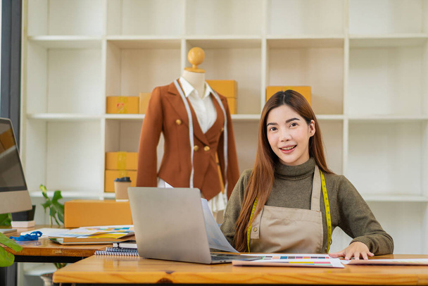 Asyalı güzel bir moda tasarımcısı, mutlu, stüdyosunda kendine gururla gülümsüyor arkasında kutular olan bir masada renkli dizüstü bilgisayarlar ve mankenlerle. sme iş fikirleri - Fotoğraf, Görsel