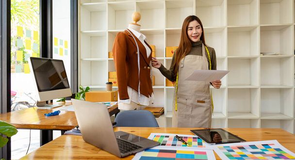 Piękna azjatycka projektantka mody, która jest szczęśliwa, uśmiecha się dumnie do siebie w swoim studio z manekinami i kolorowymi laptopami papierowymi na stole z pudełkami z tyłu. sme pomysły biznesowe - Zdjęcie, obraz