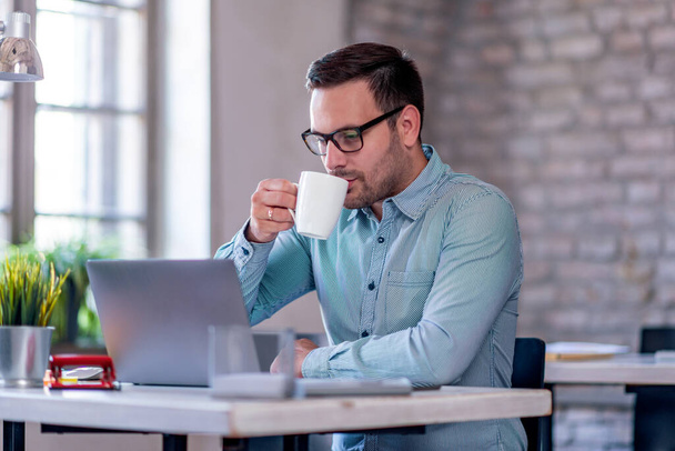 Schöner Geschäftsmann trinkt Kaffee, während er im Büro Laptop benutzt. Business, Menschen und Lifestyle-Konzept. - Foto, Bild