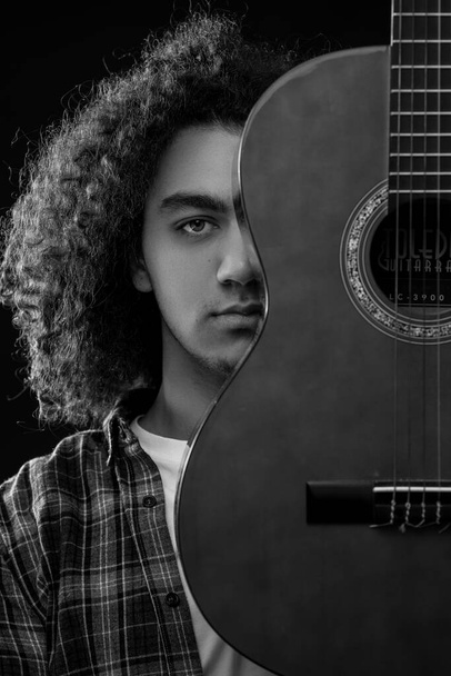 Un joven con el pelo rizado posa con una guitarra acústica delante de su cara. Foto en blanco y negro. - Foto, imagen
