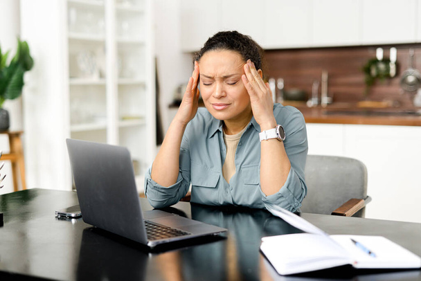 Frustrated young woman using laptop computer suffering from headache, sitting in home office and holding head with hands, feels sick and unwell - 写真・画像