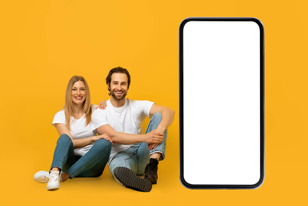 Cheerful young european couple sitting on floor with huge smartphone with empty screen - Photo, image